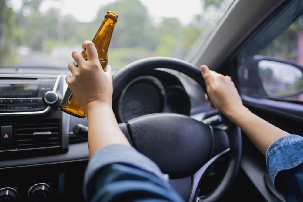 A person driving a car while holding a beer bottle in one hand.