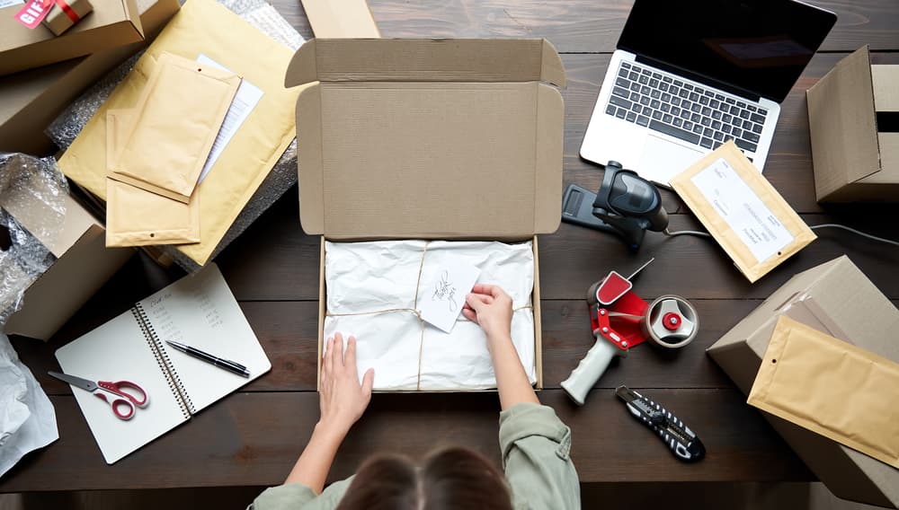 A person packing items in a box, surrounded by shipping supplies, envelopes, and a laptop.