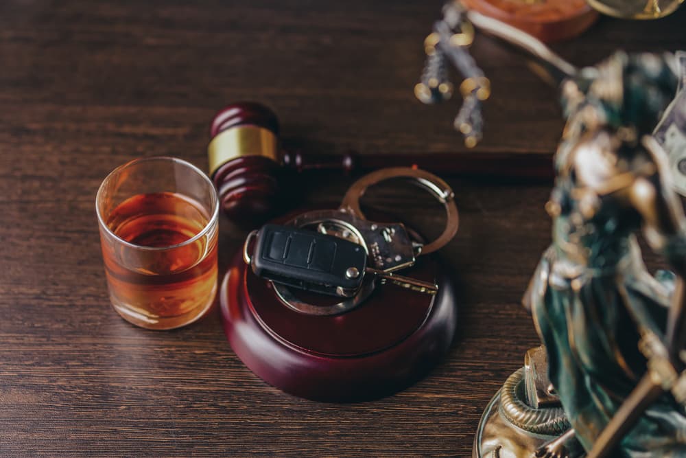 A glass of alcohol, car keys, handcuffs, and a judge's gavel on a wooden table.