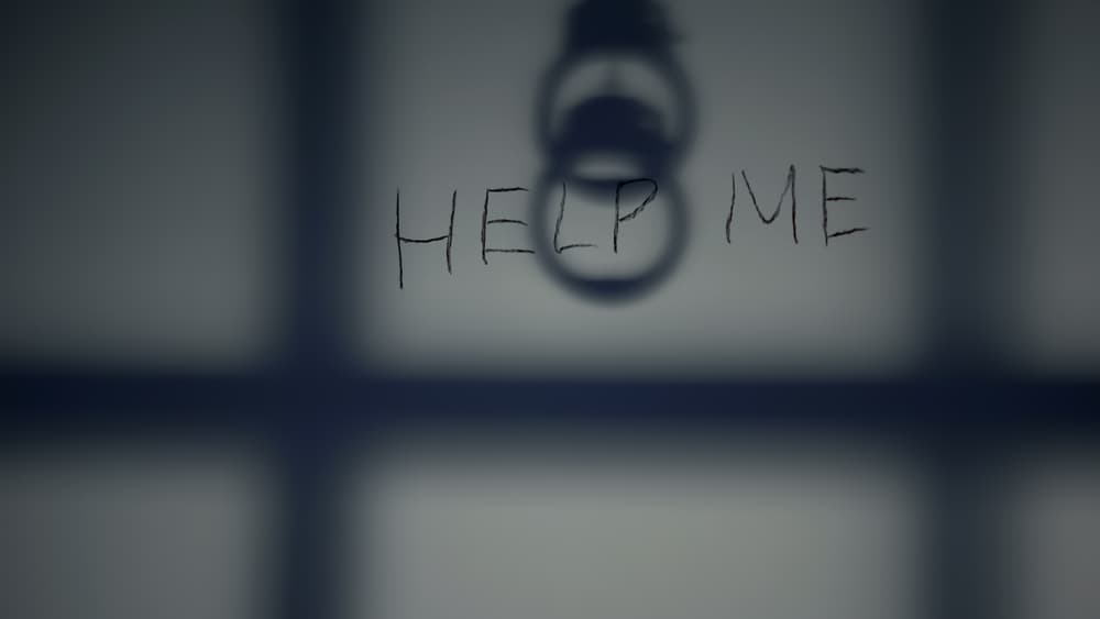 The words "HELP ME" are written on a wall, with a shadow of handcuffs above them.