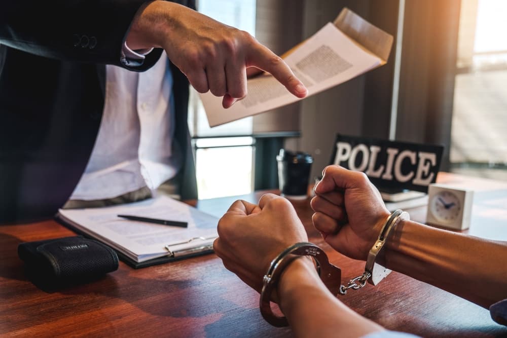 A handcuffed individual sits at a desk while a police officer points and holds a document.