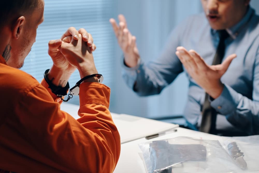 A handcuffed person in an orange jumpsuit sits across from a lawyer during a discussion.