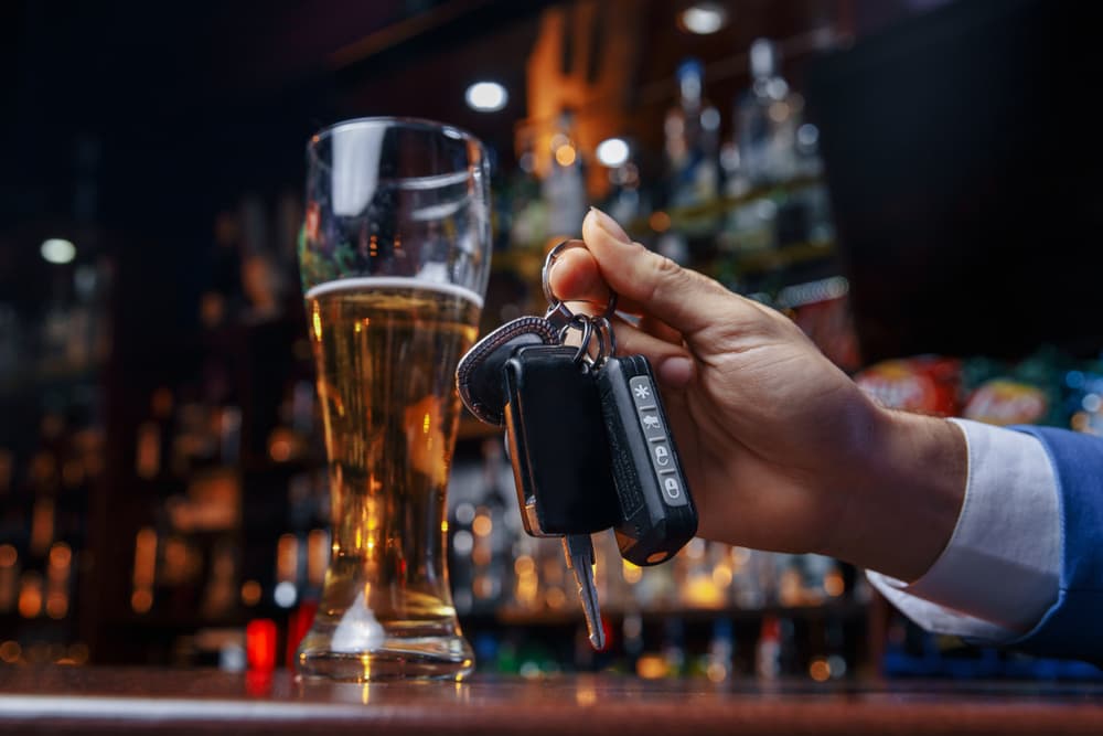 A hand holding car keys next to a glass of beer on a bar counter.