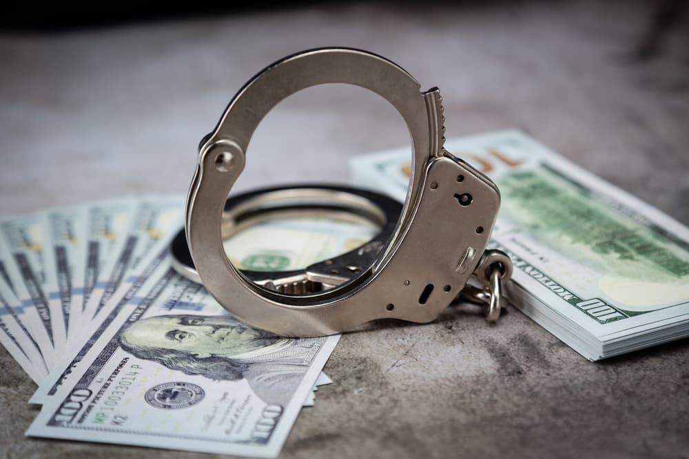 A pair of handcuffs rests on a stack of one hundred dollar bills on a table.